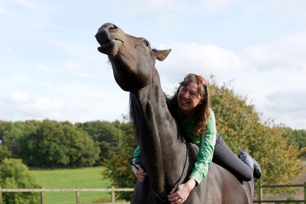 A woman lying along the back of a dark bay horse, laughing, as the horse stretches its neck up